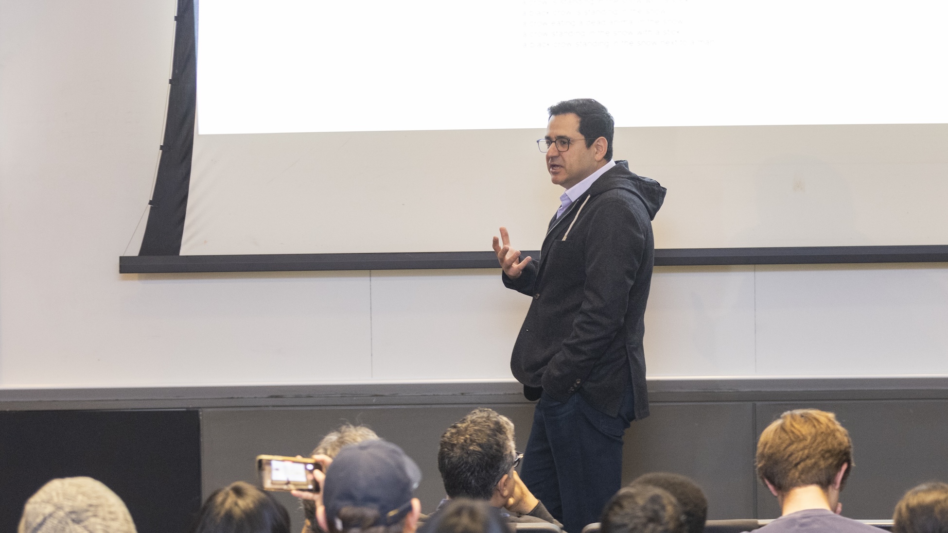 Ali Farhadi stands at the front of a lecture hall, gesturing as he speaks to an audience seated below; one attendee holds up a phone to record.