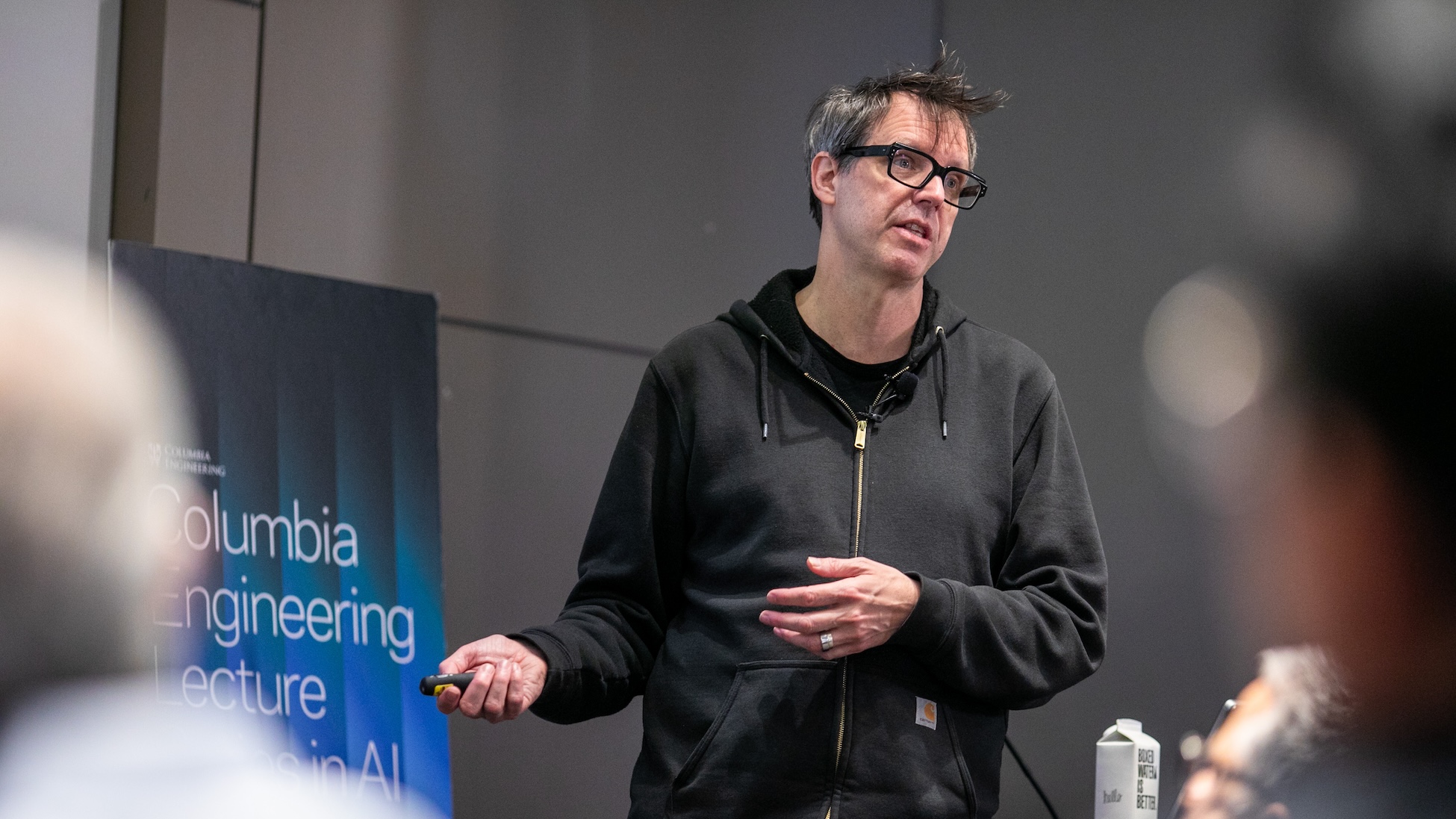 A person in a dark zip-up hoodie and glasses gives a talk indoors, holding a small presentation remote and gesturing with one hand; a blurred “Columbia Engineering” lecture sign is visible to the left and audience members are out of focus in the foreground