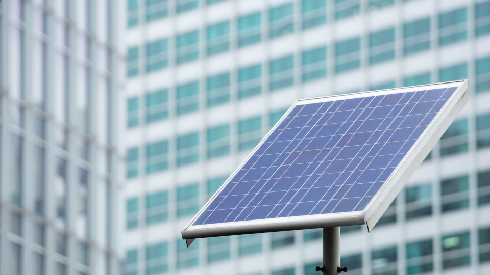 Close-up of a tilted solar panel mounted on a pole, with a blurred high-rise building of glass windows in the background.